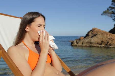 Sick tourist woman blowing nose with tissue sitting on a deck chair on a beachの写真素材