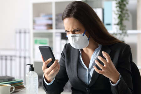 Angry executive woman with protective mask reading news on smart phone at the officeの写真素材