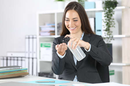 Happy executive woman applying hand sanitizer sitting on a desk at officeの写真素材