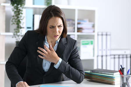 Ill executive woman suffocating and wheezing holding chest sitting at night in the officeの写真素材