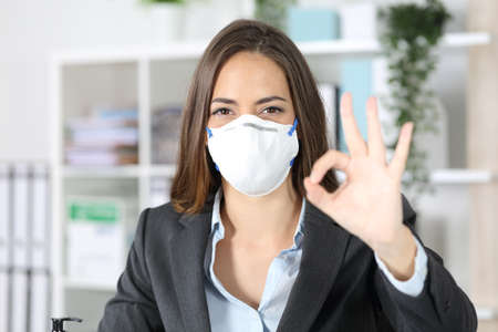 Front view of executive woman gesturing ok avoiding coronavirus looking camera with protective mask at officeの写真素材