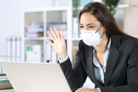 Happy executive woman looking at laptop webcam on videocall greeting avoiding covid-19 with mask sitting at the officeの写真素材