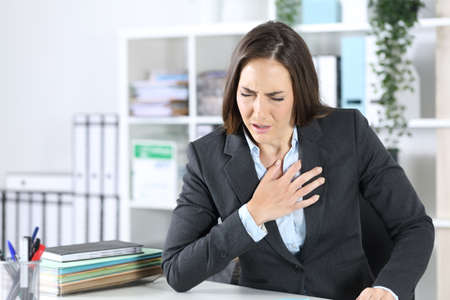 Sick executive woman suffocating holding chest sitting on a desk at officeの写真素材