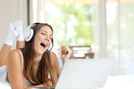 Happy girl with headphones singing listening to music on laptop lying on a bed at homeの写真素材