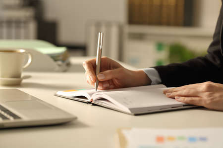 Close up of executive woman hands writing on agenda on a desk at nightの写真素材
