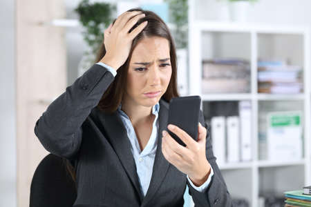 Sad worried executive woman discovers mistake on smart phone sitting on a desk at office