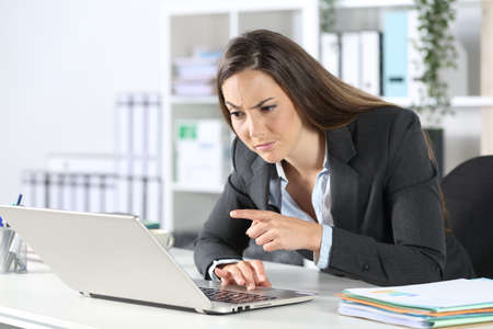 Suspicious executive woman reading and pointing on laptop sitting on a desk at the officeの写真素材