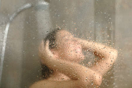 Happy woman having shower behind wet glass with drops on a bathroomの写真素材