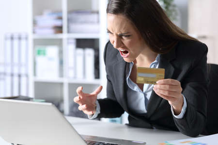 Angry executive woman pays online with credit card on laptop sitting on a desk at the officeの写真素材