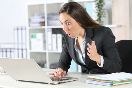 Amazed executive woman reading good news on laptop sitting on a desk at the officeの写真素材