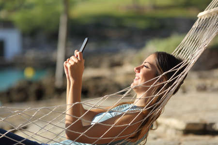 Side view portrait of a happy woman checking smart phone relaxing lying on a hammock on the beachの写真素材