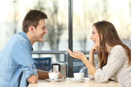 Side view portrait of a happy couple talking and flirting sitting on a table on a coffee shopの写真素材