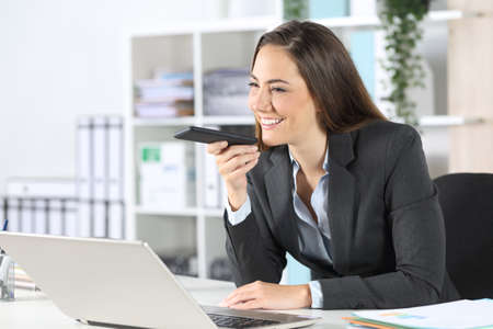 Happy executive woman using voice recognition system on smart phone sitting on a desk at the officeの写真素材