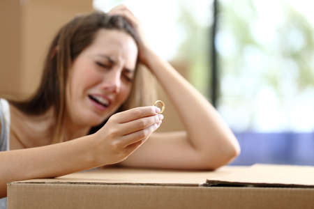 Sad wife complaining moving home after divorce holding wedding ring surrounded by boxesの写真素材
