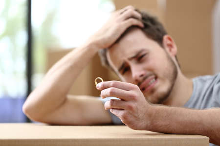 Sad husband complaining moving home after divorce and holding wedding ring surrounded by boxesの写真素材
