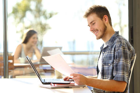 Happy student man works on laptop comparing document in a coffee shop with unfocused woman on the backgroundの写真素材