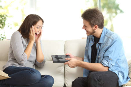 Excited surprised woman receiving gift box from man sitting on a couch in the living room at homeの写真素材