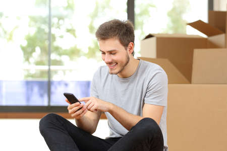 Happy man with boxes using smart phone moving home sitting on the floor surrounded by boxesの写真素材