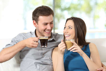 Happy couple with coffee cups looking at each other sitting in the sofa at homeの写真素材