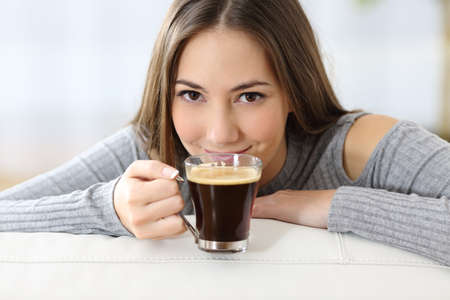 Satisfied woman smelling a coffee cup looking at camera sitting in the sofa at homeの写真素材