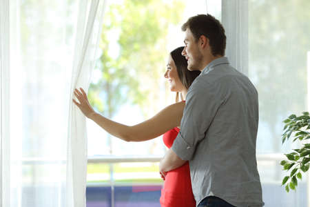 Happy couple looking through the window cuddling in the living room at homeの写真素材