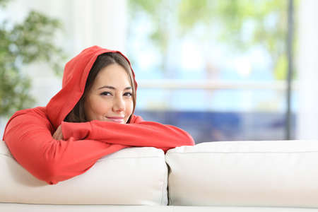 Portrait of a young woman on a red hoodie looking at camera posing sitting in the sofa at homeの写真素材