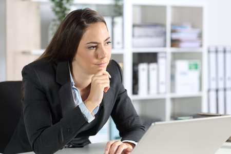 Pensive executive woman thinking looking awaywith laptop sitting on her desk in the officeの写真素材