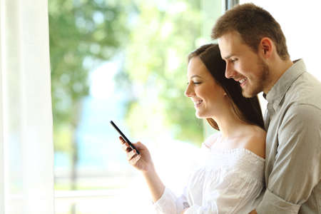 Side view of a couple checking smart phone standing next to window at homeの写真素材