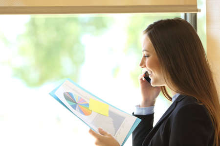 Side view of a happy executive woman checking graph report calling on smart phone standing by the window at officeの写真素材