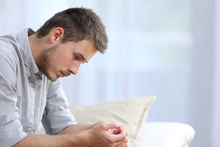 Sad man complaining looking down sitting on the sofa in the living room at homeの写真素材