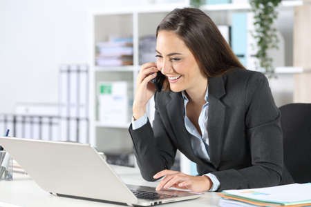 Happy executive woman calls onsmart phone using laptop sitting on a desk at the officeの写真素材
