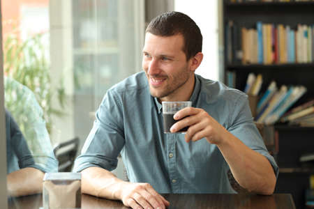 Happy man looking away throw window sitting in a coffee shop table holding glassの写真素材