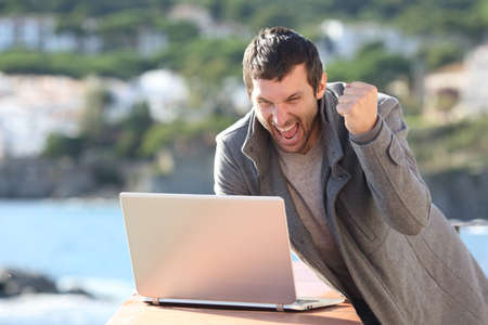 Excited man celebrating goood news on laptop on the beach in winterの写真素材