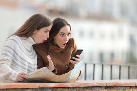 Surprised tourists women with map checking smart phone on a balcony in winterの写真素材