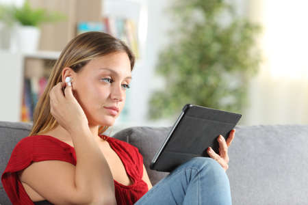 Serious woman listening music with earbuds on a tablet sitting on the couch at homeの写真素材