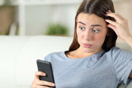 Overwhelmed girl discovering mistake on smart phone sitting on a couch in the living room at homeの写真素材