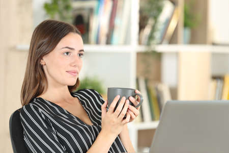 Relaxed woman holding coffee mug contemplating sitting on a chair at homeの写真素材