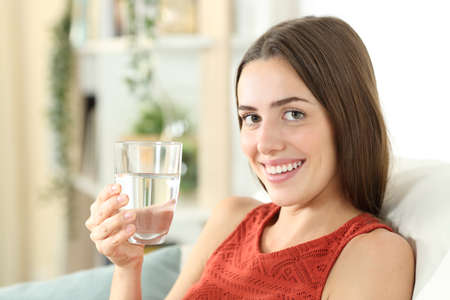 Happy woman is holding a mineral water glass sitting on a couch in the living room at homeの写真素材