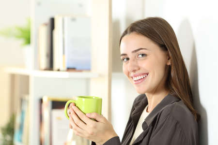 Happy woman holding green coffee cup looks at camera standing at homeの写真素材