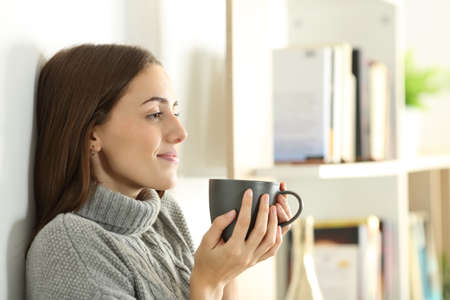 Profile of a relaxed woman looks away in winter holding coffee mug standing at homeの写真素材