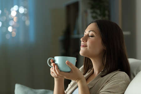 satisfied woman relaxing smelling coffee in the night sitting on a couch in the living room at homeの写真素材