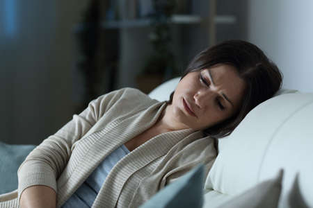 Sad woman looking down sitting on a couch in the living room at home in the nightの写真素材
