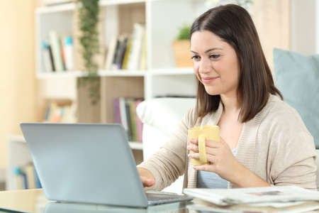 Satisfied woman drinking coffee using a laptop on a desk at homeの写真素材