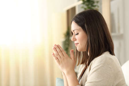 Side view of a christian religious woman praying sitting on a couch in the living room at home with window light in the backgroundの写真素材