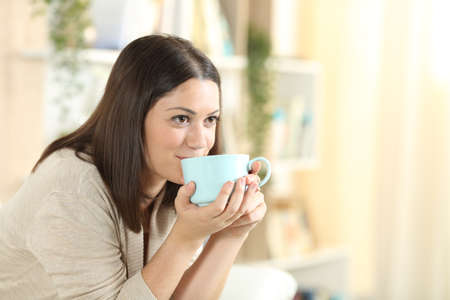Happy woman drinking coffee sitting on a sofa looking away in the living room at homeの写真素材