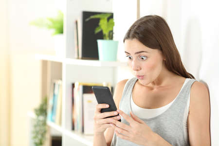 Surprised woman checks mobile phone content standing at homeの写真素材