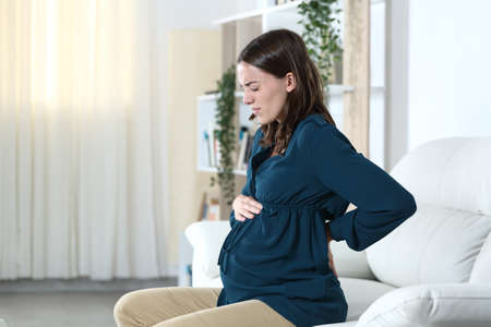 Pregnant woman with back ache complaining sitting on a sofa in the living room at homeの写真素材