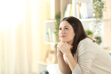 Pensive woman looks up dreaming sitting on a sofa at homeの写真素材