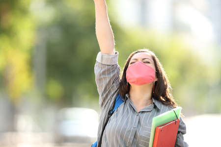 Front view portrait of an excited student wearing mask raising arm in the street celebrating success