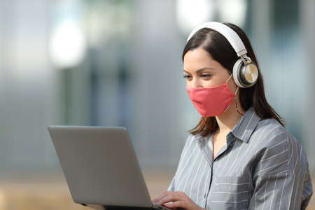 Happy woman with mask using laptop and headphones sitting in the street